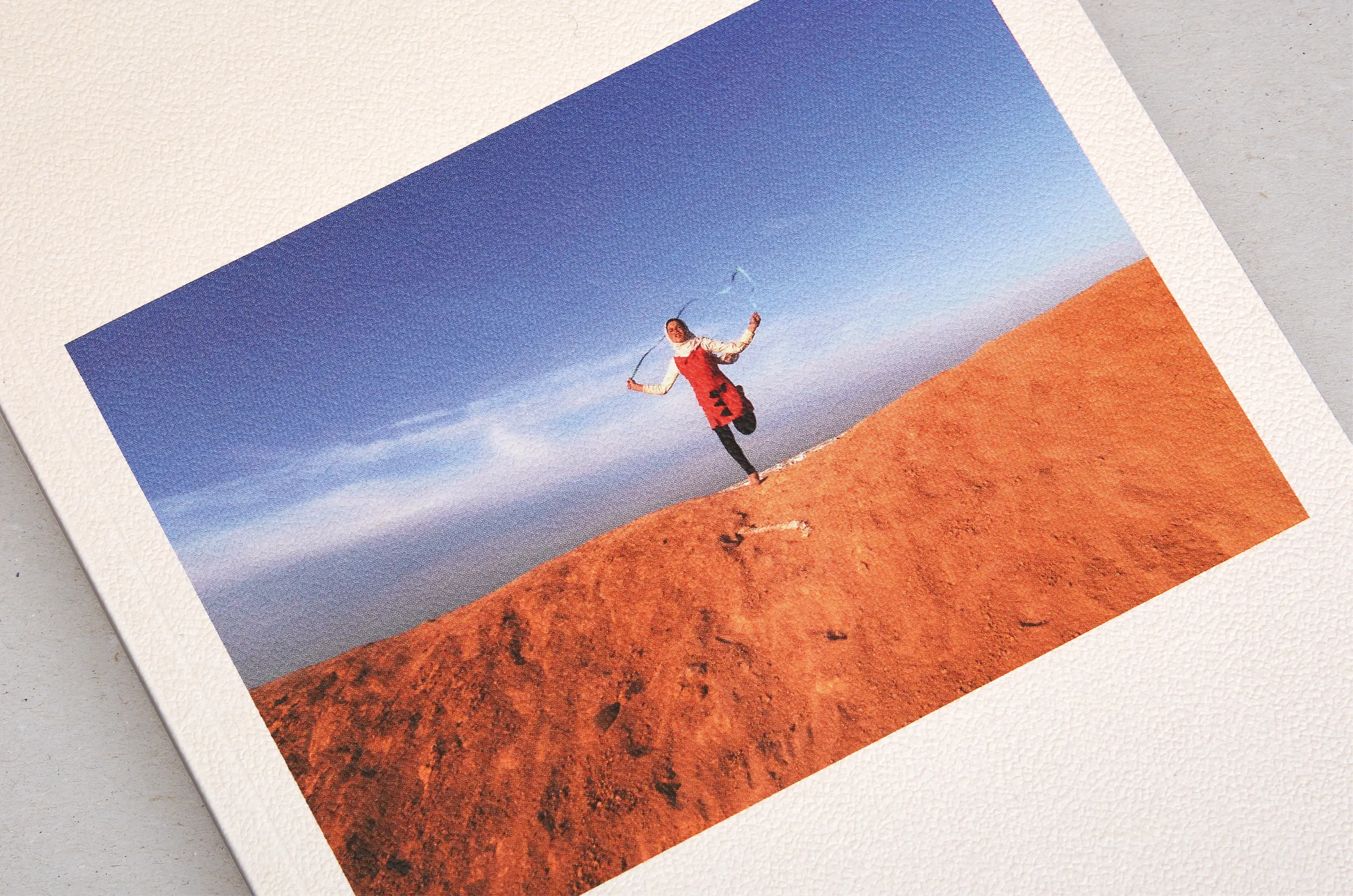 Exhibition print of a child's photograph — a figure skipping on red desert ground under a blue sky